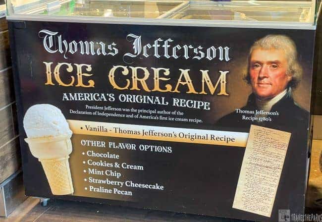 a sign for "Thomas Jefferson Ice Cream" displayed at a counter or stall. The sign prominently features the words "Thomas Jefferson Ice Cream" in large, ornate lettering, with the phrase "America's Original Recipe" below it. To the right side of the sign is a portrait of Thomas Jefferson, with a note stating "Thomas Jefferson’s Recipe 1780s." Below the main title, there's a text box for the flavor "Vanilla - Thomas Jefferson's Original Recipe" and a list of other flavor options including Chocolate, Cookies & Cream, Mint Chip, Strawberry Cheesecake, and Praline Pecan. An image of a vanilla ice cream cone is shown on the left side. A section of historical handwritten text is depicted on the lower right side of the sign.