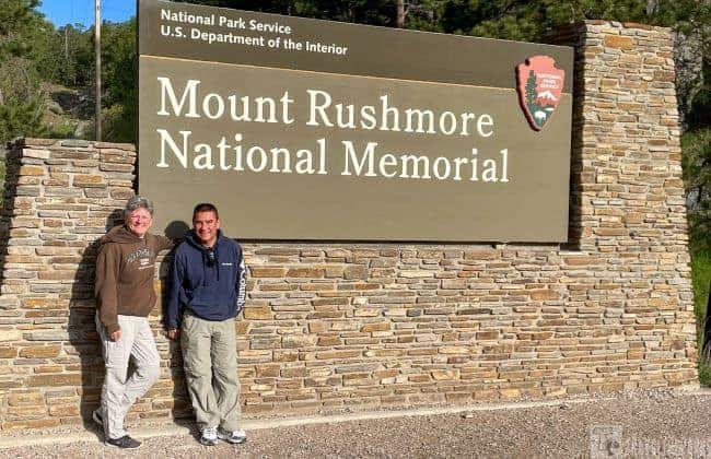 Two people stand in front of the Mount Rushmore National Memorial sign.