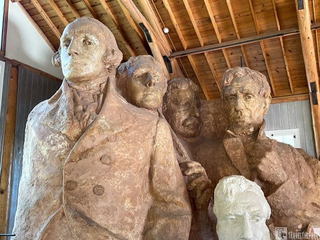 Indoor sculpture model of Mount Rushmore showing busts of figures in a tan material with a wooden ceiling backdrop.