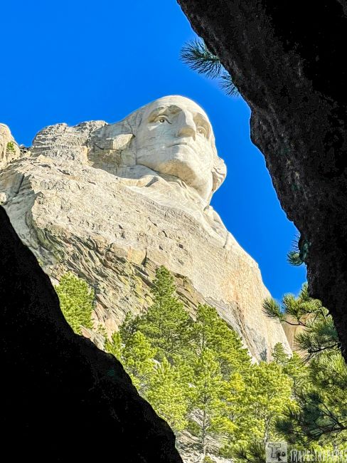 A carved face of George Washington on Mount Rushmore framed by rocky outcrops and trees, under a bright blue sky.