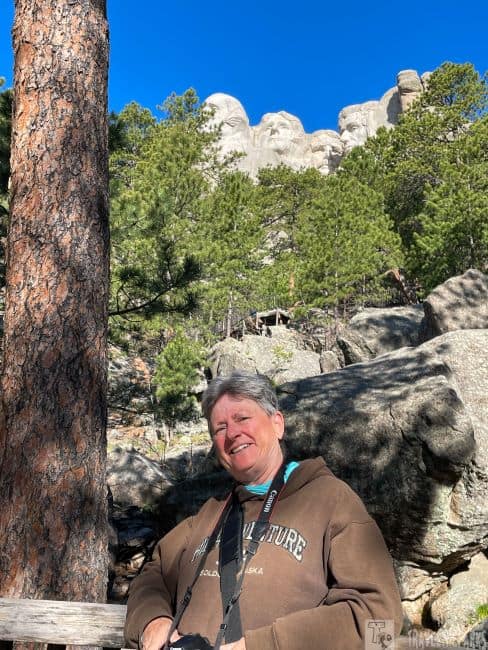 A person smiling in front of a natural setting with pine trees and Mount Rushmore in the background.