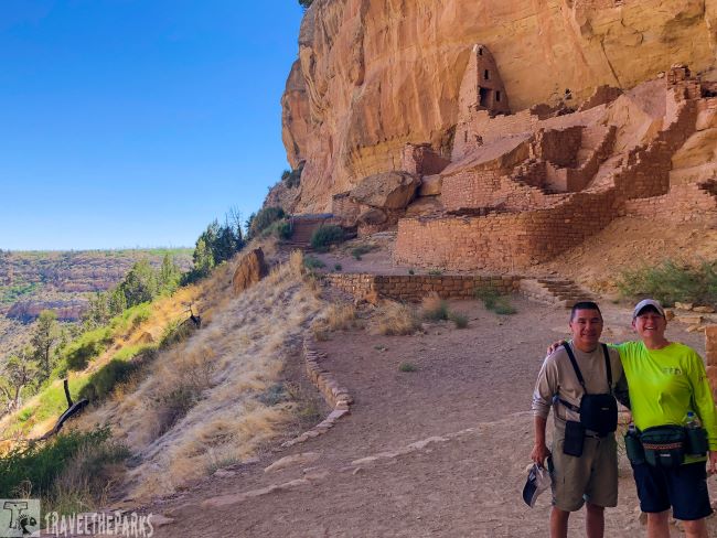 Two people stand on a path by ancient stone ruins of Longhouse set into a large cliff.