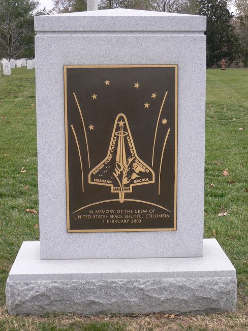 A memorial stone with a bronze plaque commemorating the U.S. Space Shuttle Columbia crew, set in a grassy cemetery.