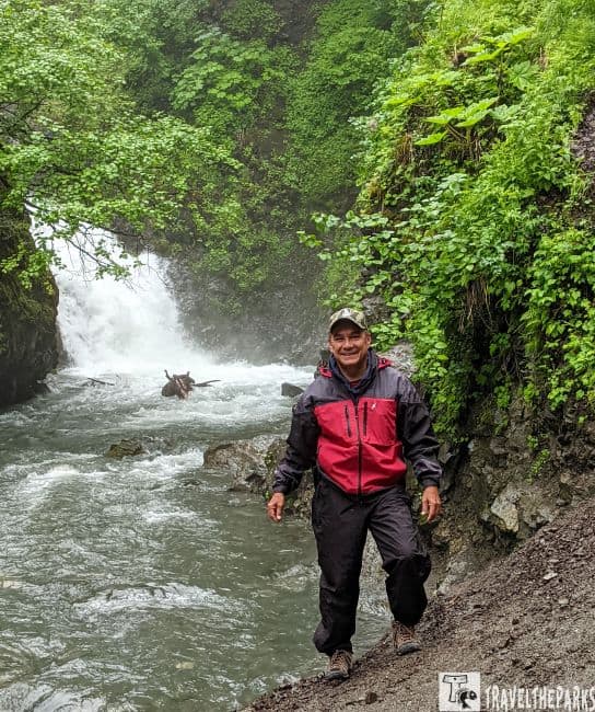 a person standing in front of a waterfall, surrounded by lush, green vegetation. The waterfall, nestled between steep, rocky slopes, cascades into a flowing river. The person is wearing a red and black waterproof jacket and dark pants, with a cap on their head, and stands on a rocky, slightly muddy riverbank. The foliage on the banks is dense, with various shades of green creating a vibrant natural setting. The sky above is not visible, suggesting a dense canopy or a close-up perspective.
